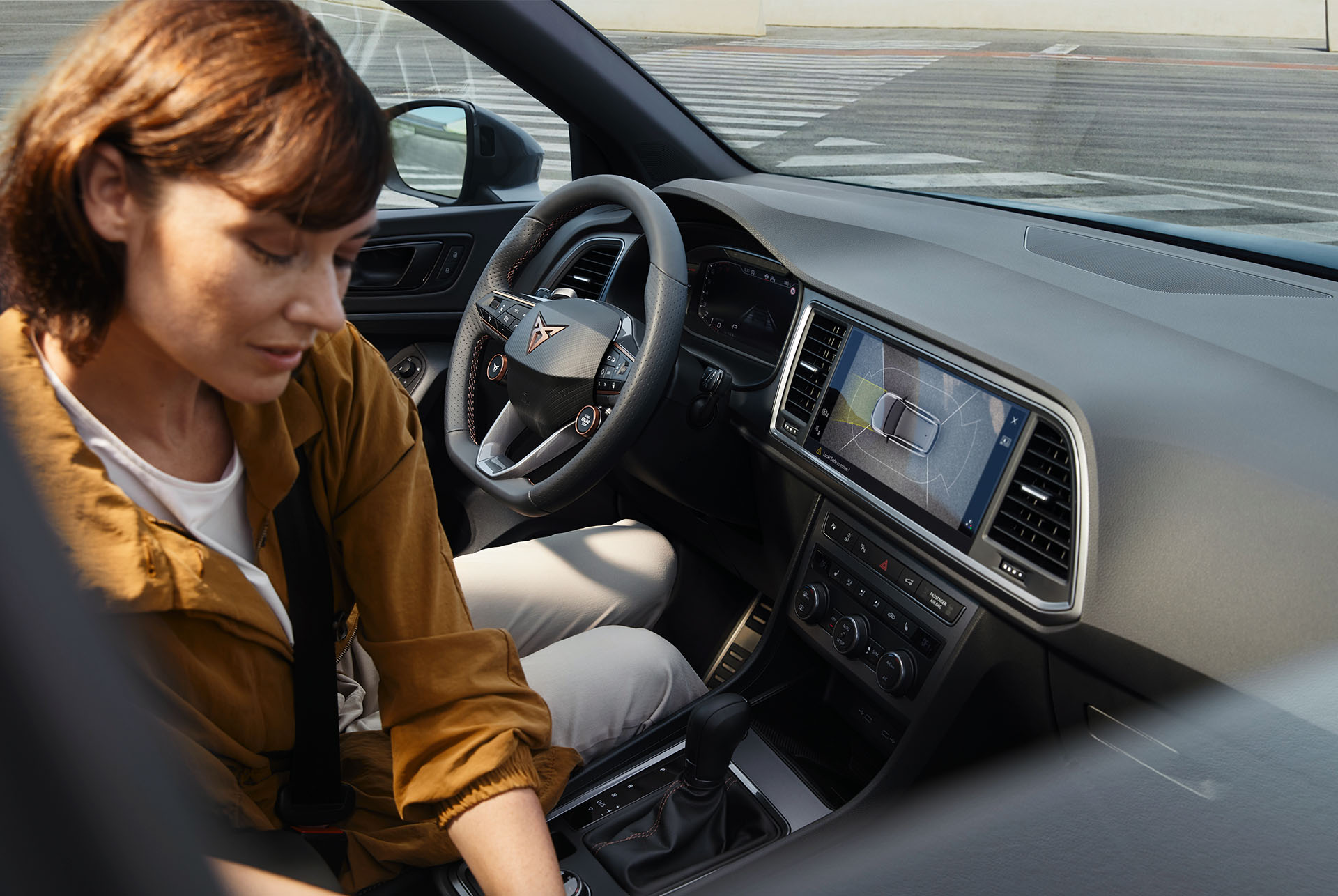 The interior of the cupra ateca highlighting advanced technology features. The dashboard displays a large infotainment screen with navigation and the steering wheel with satellite buttons has integrated controls. A woman is seated inside. 