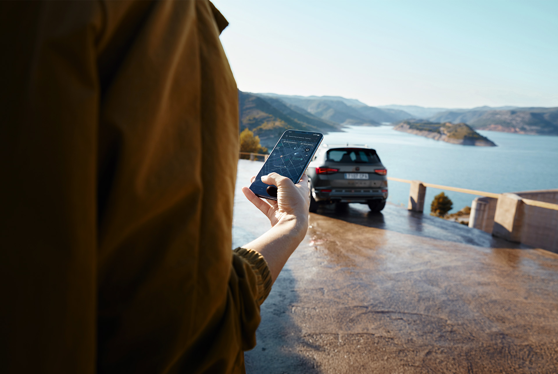 A person holding a smartphone with the CUPRA Ateca in the background, parked near a scenic view of mountains and water, symbolizing a connected world.