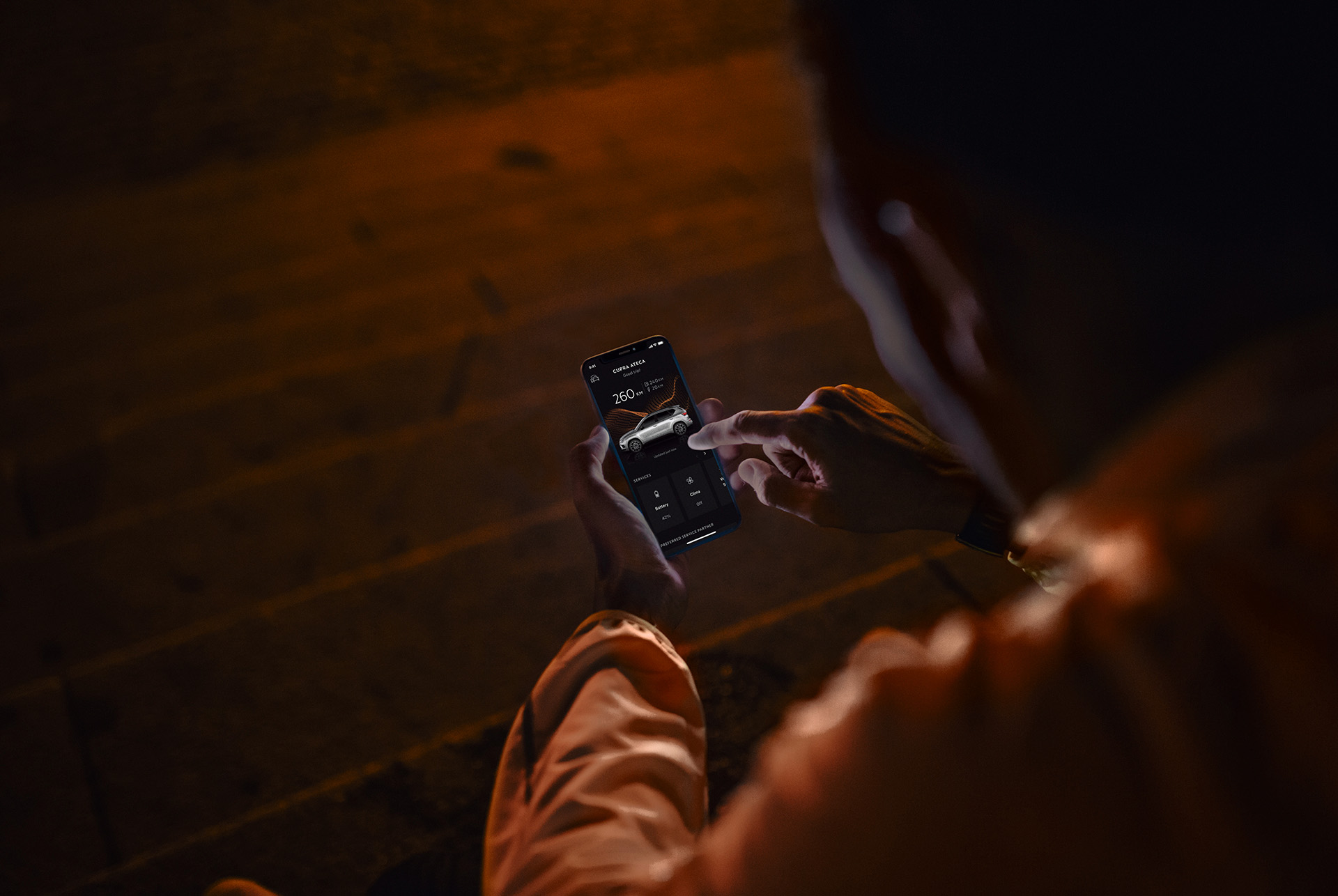 CUPRA CONNECT online services incorporates a range of solutions to answer to the challenges of modern life. A person sitting on stairs at night, using a smartphone illuminated by the screen.